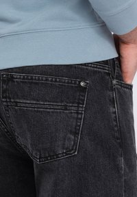 Jeans de mezclilla gris oscuro con una textura suave, que cuentan con un bolsillo trasero y detalles de remaches metálicos, combinados con una sudadera azul clara.