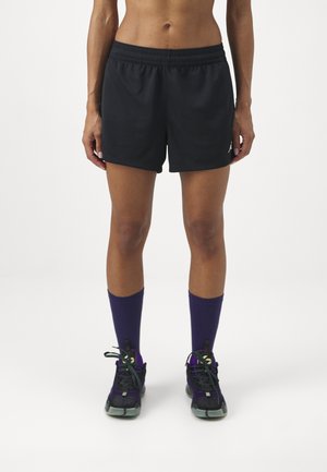 Schwarze Sportshorts mit einem elastischen Bund, die eine glatte Oberfläche und ein kleines Logo aufweisen. Kombiniert mit lila Kniestrümpfen und schwarzen Sneakers.