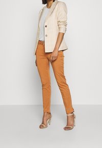 Blazer à carreaux beige, t-shirt gris, pantalon ajusté orange et sandales à talons hauts nude. Le blazer présente des revers crantés et deux boutons.