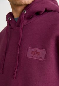 Hoodie bordeaux en tissu doux, avec un patch logo sur le devant et des cordons de serrage au niveau de la capuche avec des œillets métalliques.