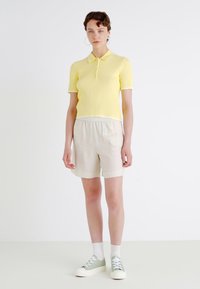 Polo shirt giallo a righe con colletto bianco, abbinato a pantaloni corti beige chiaro e sneakers verdi. Il modello è in piedi contro uno sfondo semplice.