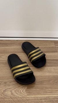 Chinelos pretos de borracha com três riscas horizontais douradas, apresentando um design de ponta aberta e sola plana.