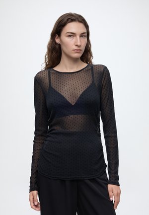 Jeune femme aux longs cheveux ondulés portant un haut noir transparent à manches longues avec de petits pois et un pantalon noir, debout devant un fond uni.