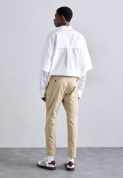 Maglietta bianca oversize con spalle cadute, chinos beige e sneakers con motivo bianco, verde e bordeaux. Sfondo grigio uniforme.