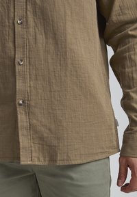 Chemise boutonnée marron clair en tissu texturé, avec des boutons argentés et un ourlet arrondi, portée avec un pantalon vert olive.