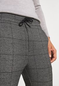 Szare spodnie w kratę z sznurkowym pasem i teksturowaną tkaniną, podkreślające krzyżujące się jasne i ciemne linie wzoru.