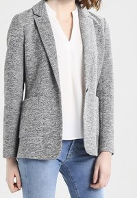 Veste grise texturée avec un col cranté, deux poches avant et une coupe ajustée, portée sur un chemisier blanc et un jean bleu clair.