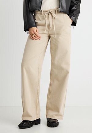 Μπεζ παντελόνι ευρέος ποδιού από τζιν με κορδόνι στη μέση, ορατά ραφές και λεία υφή, συνδυασμένο με μαύρα πλατφόρμα παπούτσια.