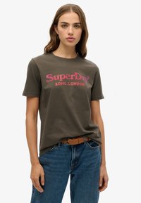 Camiseta de algodón verde oliva con cuello redondo y mangas cortas, que presenta el texto rosa "SuperDry SOHO LONDRES" en una fuente en negrita.