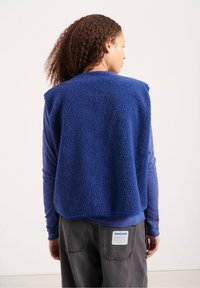 Mėlynas vilnos liemenė su tekstūruota, puria išore, su atviru nugara. Nešiojama ant mėlynos ilgomis rankovėmis marškinėlių ir pilkų kelnių.