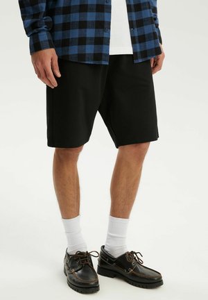 Mężczyzna noszący czarne spodenki do kolan, białe skarpetki, czarne buty żeglarskie, białą koszulę oraz niebiesko-czarną kratowaną koszulę na wierzch.