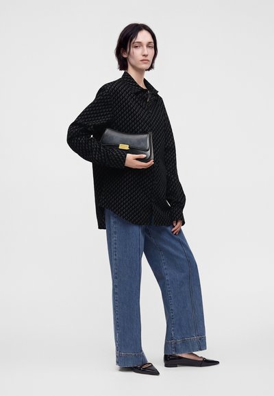 Femme debout tenant une pochette noire, portant une chemise noire à motifs oversize, un jean bleu à jambes larges et des ballerines noires pointues sur fond uni.