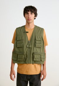 Πράσινο γιλέκο εργαλείου με πολλαπλές μπροστινές τσέπες, κλείσιμο με φερμουάρ και χαλαρή εφαρμογή, φορεμένο πάνω από ένα μουσταρδί μπλουζάκι. Απλός σχεδιασμός με ελάχιστα αξεσουάρ.