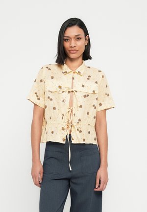 Blusa de manga corta color amarillo claro con estampado floral, detalle de anudado en el frente, tela texturizada y diseño peplum. Combinada con pantalones gris oscuro.