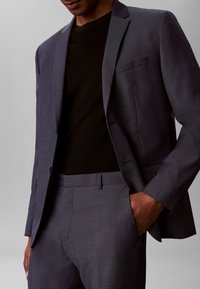 Homme portant un costume gris foncé avec pantalon assorti et une chemise noire, une main dans la poche devant un fond gris uni.