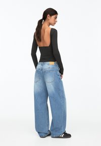 Schwarzes Langarmoberteil mit tiefem Rückenausschnitt, kombiniert mit übergroßen hellblauen Denim-Jeans. Die Jeans verfügen über zwei Gesäßtaschen und ein braunes Etikett.