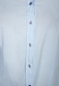 Light blue button-up shirt featuring fabric texture, six buttons, and contrasting button details. Simple design with a classic collar style.