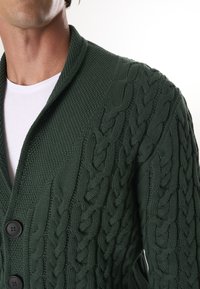 Mørkegrøn kabelstrikket cardigan med sjalskrave, tekstureret mønster og sorte knapper. Materialet virker tykt og behageligt.