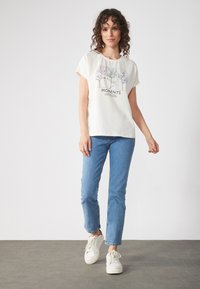Hea kreemjas lühikeste varrukatega T-särk lillelise graafikaga ja tekstiga "MOMENTS", mis on kombineeritud helesiniste denim pükstega ja valgete spordijalatsitega.