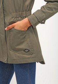 Olivově zelená bunda s tahem v pase, vybavená velkými předními kapsami a dekorativním logem na dolní kapse. Lehce průsvitný materiál.