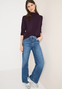 Lila langärmliges Oberteil mit hohem Kragen, kombiniert mit weit geschnittenen blauen Jeans. Das Modell trägt silberne Sneakers und steht vor einem neutralen Hintergrund.