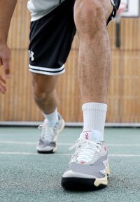 Szürke és fehér sportcipők piros kiegészítőkkel, párnázott kialakítással, texturált anyaggal és fekete gumi talppal, fehér zoknival viselve.