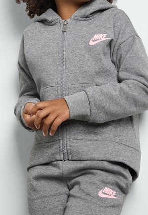 Kind trägt grauen Nike-Trainingsanzug mit pinken Logos, die Hände verschränkt, steht vor einer hellen Wand.
