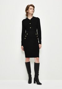 Schwarzes geripptes Strickkleid mit rundem Ausschnitt, Knopfleiste, tailliertem Schnitt und goldenen Knopf-Akzenten an den Hüften, kombiniert mit schwarzen kniehohen Stiefeln.