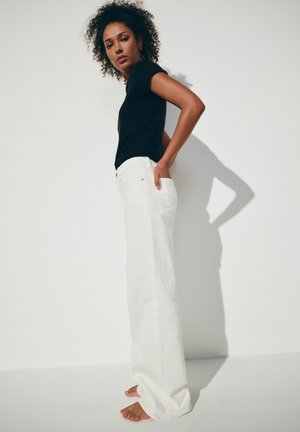 Femeie cu păr creț, purtând un top negru cu mâneci scurte și pantaloni albi, largi și cu talie înaltă, stând desculță în fața unui perete alb.