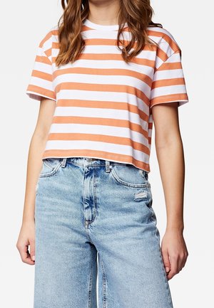 Henkilö pukeutuneena lyhennettyyn oranssi-valkoiseen raidalliseen t-paitaan ja vaaleansinisiin korkeavyötäröisiin farkkuihin, joissa on pieni repeämä.