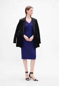 Mørkeblå tettsittende kjole med V-hals, lagdelt med en svart oversized blazer. Svart sandaler med hæl og stropper fullfører antrekket.