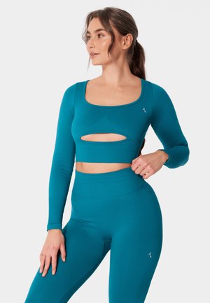 Kobieta z długimi brązowymi włosami związanymi w kucyk, ubrana w turkusową bluzkę typu crop top z długim rękawem i wycięciem na klatce piersiowej oraz dopasowane legginsy z wysokim stanem na białym tle.