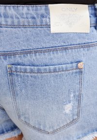 Pantalones cortos de mezclilla en azul claro con dobladillo deshilachado. Presentan un bolsillo trasero, costuras visibles y una etiqueta en la pretina con el nombre de la marca.