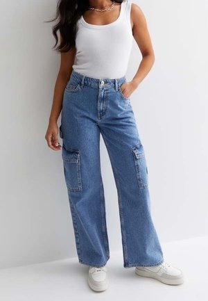 Vrouw met een hoge taille blauwe denim cargobroek, witte tanktop, witte sneakers en een zilveren ketting halsketting, staand tegen een witte achtergrond.