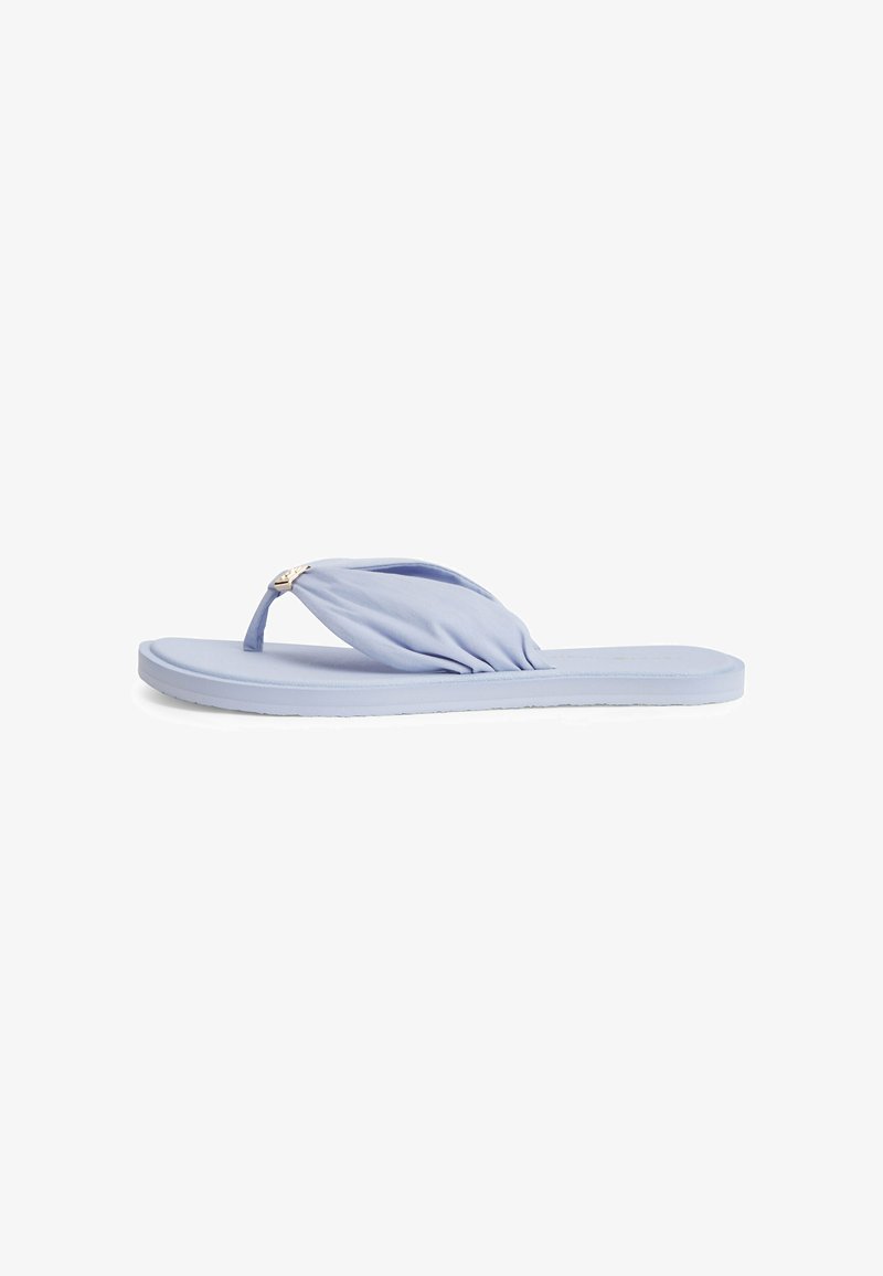 Sandale plate bleu clair avec bride en tissu rembourrée et petit détail de logo métallique sur fond blanc.