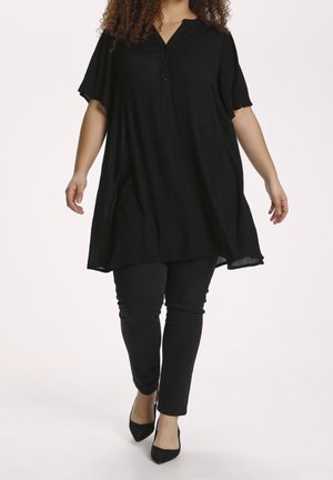 Femme marchant, portant une tunique noire ample à manches courtes, un pantalon noir ajusté, et des talons noirs à bout pointu sur fond blanc.