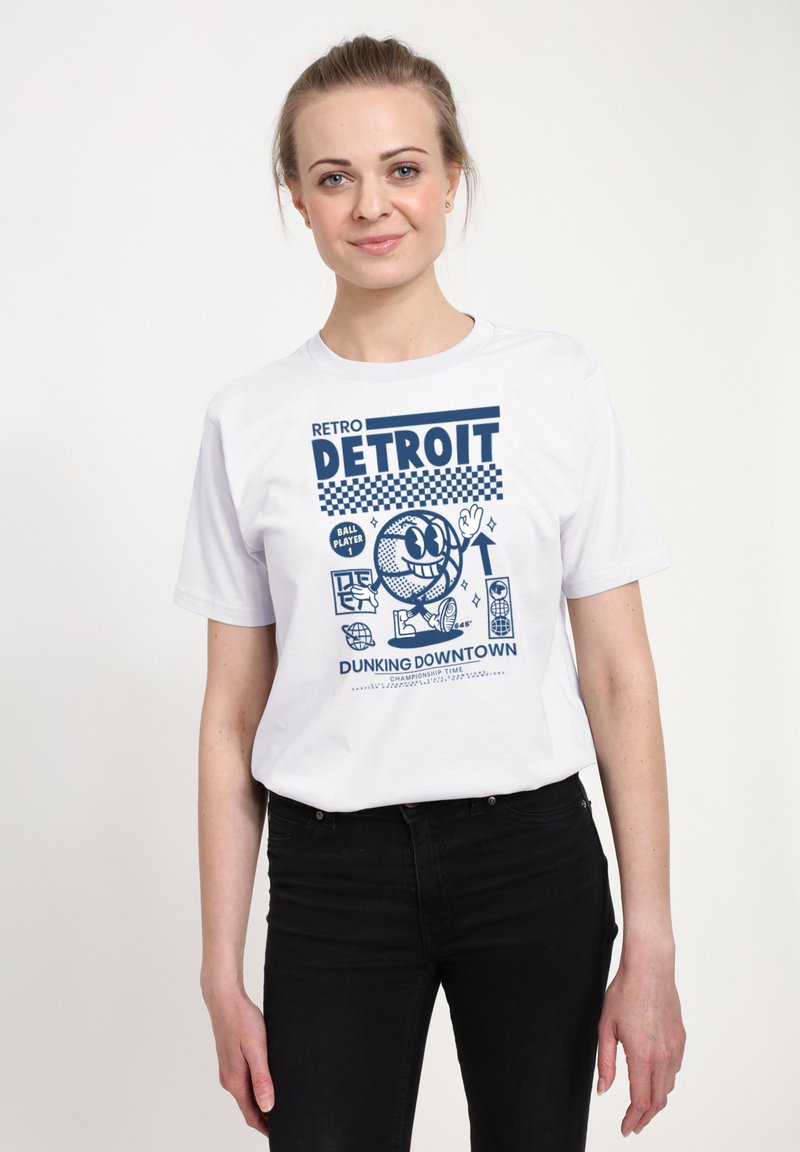 Mladá žena oblečená v bílém tričku s modrým retro grafickým motivem basketbalu Detroit a černými kalhotami, stojící proti jednoduchému bílému pozadí.