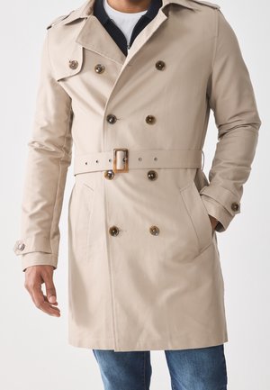 Trench-coat beige à double boutonnage avec ceinture et détails de boutons porté sur une chemise blanche et un jean bleu, une main dans la poche.