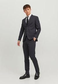 Completo grigio scuro con vestibilità slim, blazer a un bottone, pantaloni abbinati e una camicia bianca. Abbinato a scarpe in pelle nere.