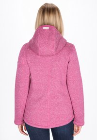 Chaqueta de forro polar rosa con capucha. Textura suave, detalles de costura en la parte posterior y dobladillo ligeramente curvado. Combinada con jeans azules.