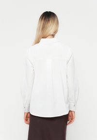 Vit blus med lång ärm och en veck bak, knäppte manschetter och krage. Materialet ser slätt ut med en strukturerad design.