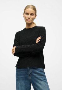 Vrouw met blond haar in een paardenstaart, draagt kleine oorbellen, een zwart langharig shirt en een blauwe spijkerbroek, met haar armen gekruist, tegen een witte achtergrond.