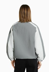 Šedá lehká bunda s bílými akcenty, volný střih, elastické manžety a šňůrkový lem. Má hladkou texturu a geometrický design.