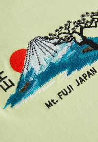 Světlá zelená tkanina s barevným vyšívaným vzorem hory Fuji, červeného slunce a černého stromu, nápis zní "Mt. FUJI JAPONSKO."