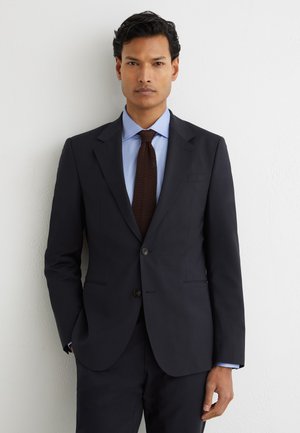 Mann mit dunklem Haar, der einen schwarzen Anzug, blaues Hemd und dunkelbraune Krawatte trägt, steht vor einer weißen Wand, die Hände teilweise in den Taschen.