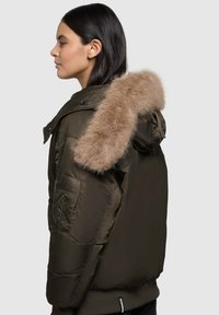 Alyvuogių žalios spalvos pūkinė striukė su dirbtinio kailio gaubtu, užtrauktuku užsegama šoninė kišenė, siūtos tekstūros ir trupinėjančiu hemu.