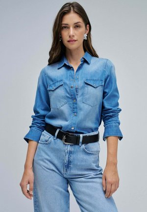 Camisa de mezclilla azul claro con botones de presión, dos bolsillos en el pecho y mangas enrolladas, combinada con jeans azul claro y un cinturón negro.