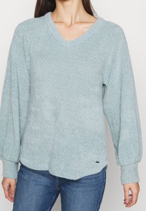 Maglione lavorato a maglia azzurro chiaro con scollo a V, maniche lunghe a sbuffo, superficie testurizzata e orlo leggermente curvo. Piccola etichetta con logo di lato.