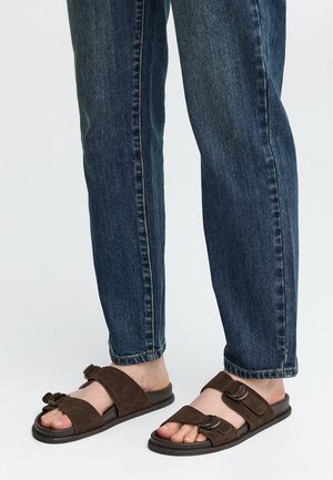 Füße, die dunkelbraune Wildleder-Sandalen mit Schnallen tragen, kombiniert mit blauen Jeans vor einem schlichten hellen Hintergrund.