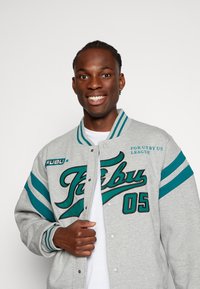 Gray varsity jacket with teal stripes and embroidered text. Features a button closure, ribbed cuffs, and a relaxed fit design.